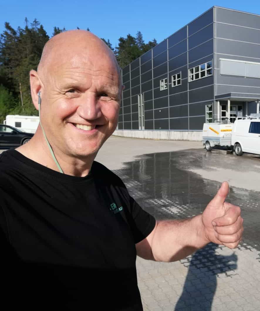 Smilende mann tar selfie foran industribygg og hvit arbeidsbil med tilhenger.
