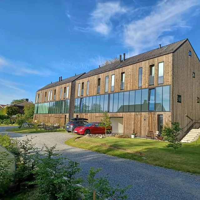 Rekkehus med trefasade og store glassflater i solskinn, biler parkert foran.